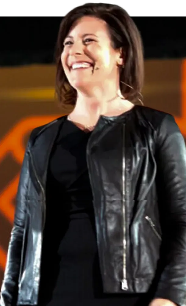 Woman in black dress and leather jacket speaking on stage.